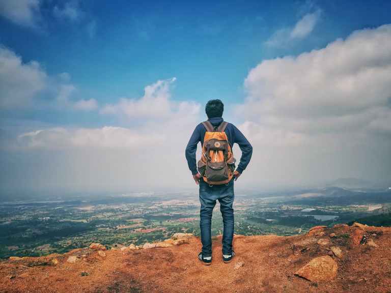 man in blue dress shirt and blue jeans and orange backpack standing on mountain cliff looking at town under blue sky and white clouds